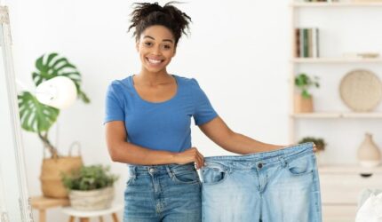 Wanita kulit hitam ramping yang bahagia menunjukkan jeans kebesaran lama setelah diet penurunan berat badan yang sukses